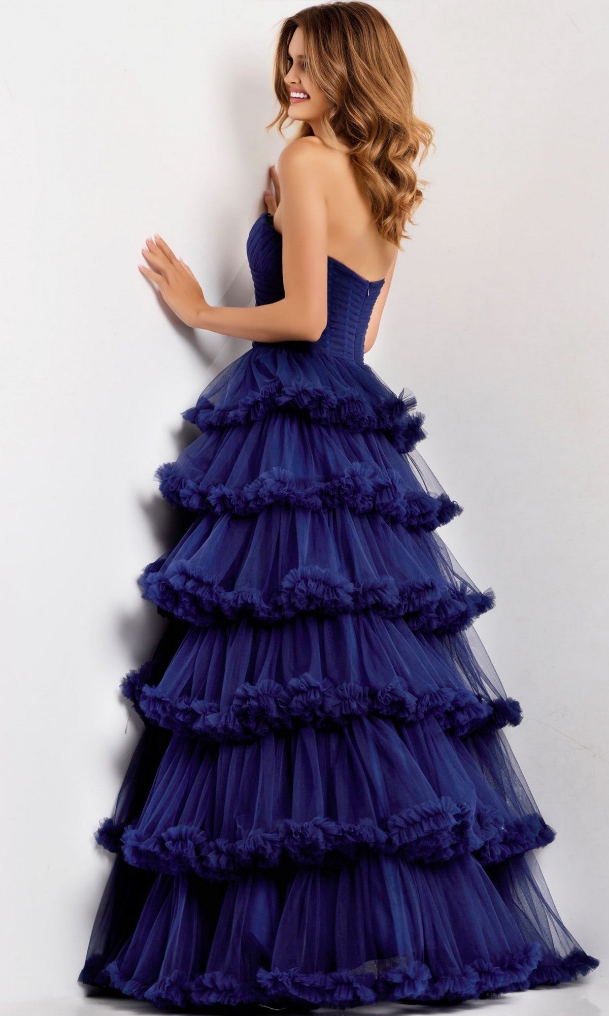 A woman poses sideways with her hand on the wall, wearing the Long Prom Dress: Jovani 47139. The dark blue strapless gown features elegant, tiered ruffles for a stunning silhouette against a light background.